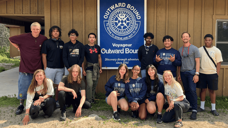 The image shows a group of people posing in front of a building with an "Outward Bound" sign. There are both adults and teenagers in the group, and they are all smiling. The setting appears to be outdoors, possibly at a camp or educational facility.